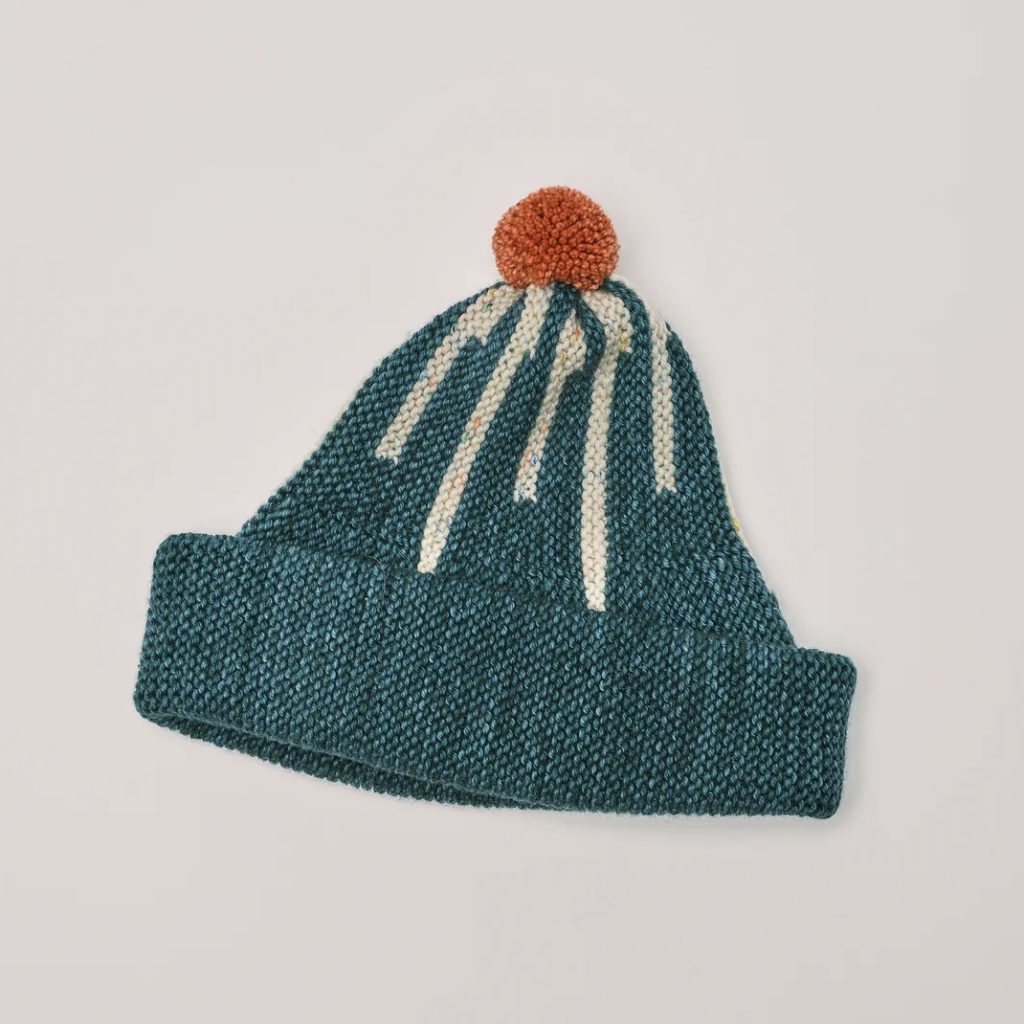 A hand knit cap with an abstract star pattern in a green-blue with an orange pom pom